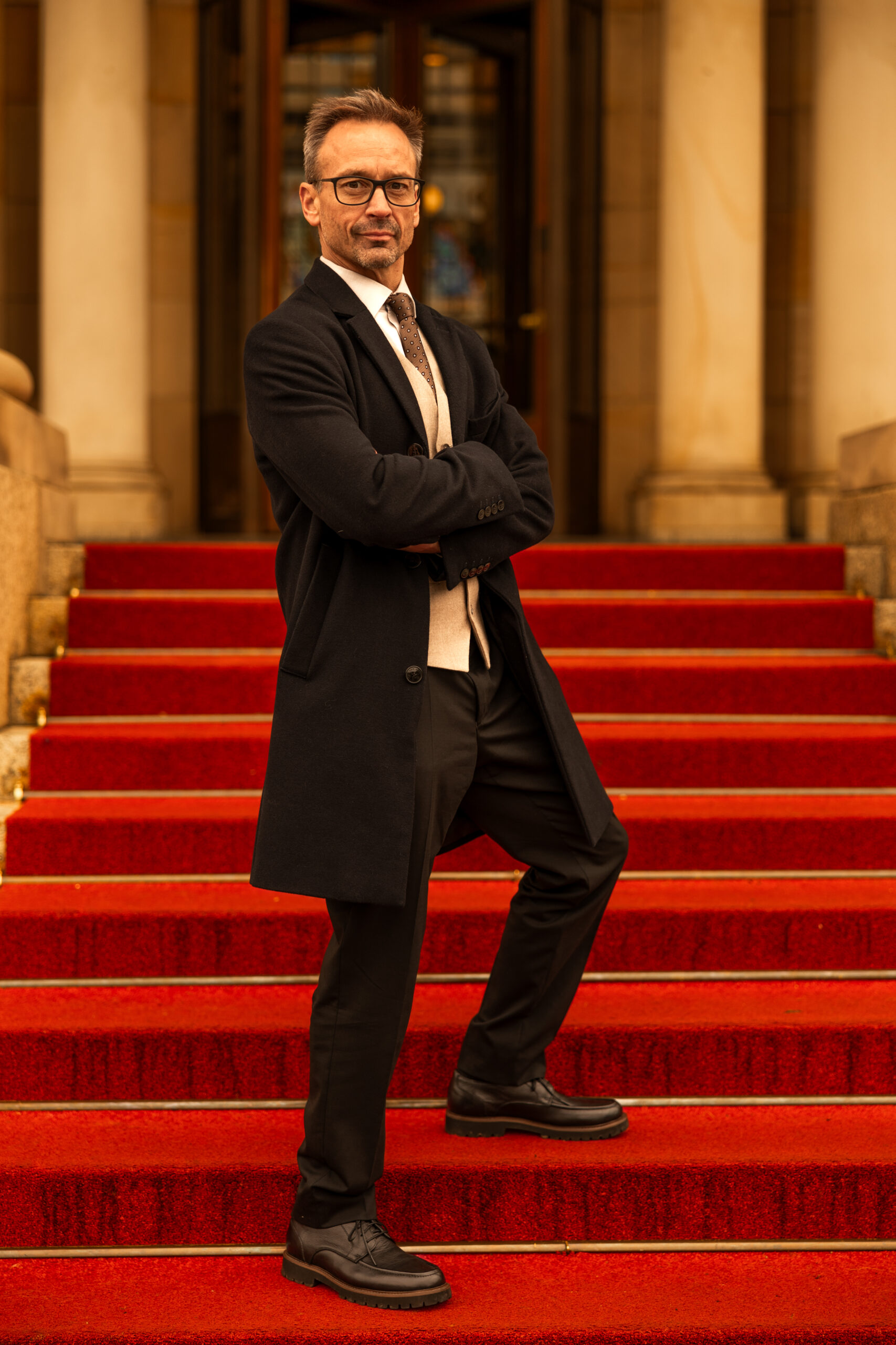 John Caulwell standing with arms crossed on red carpeted stairs