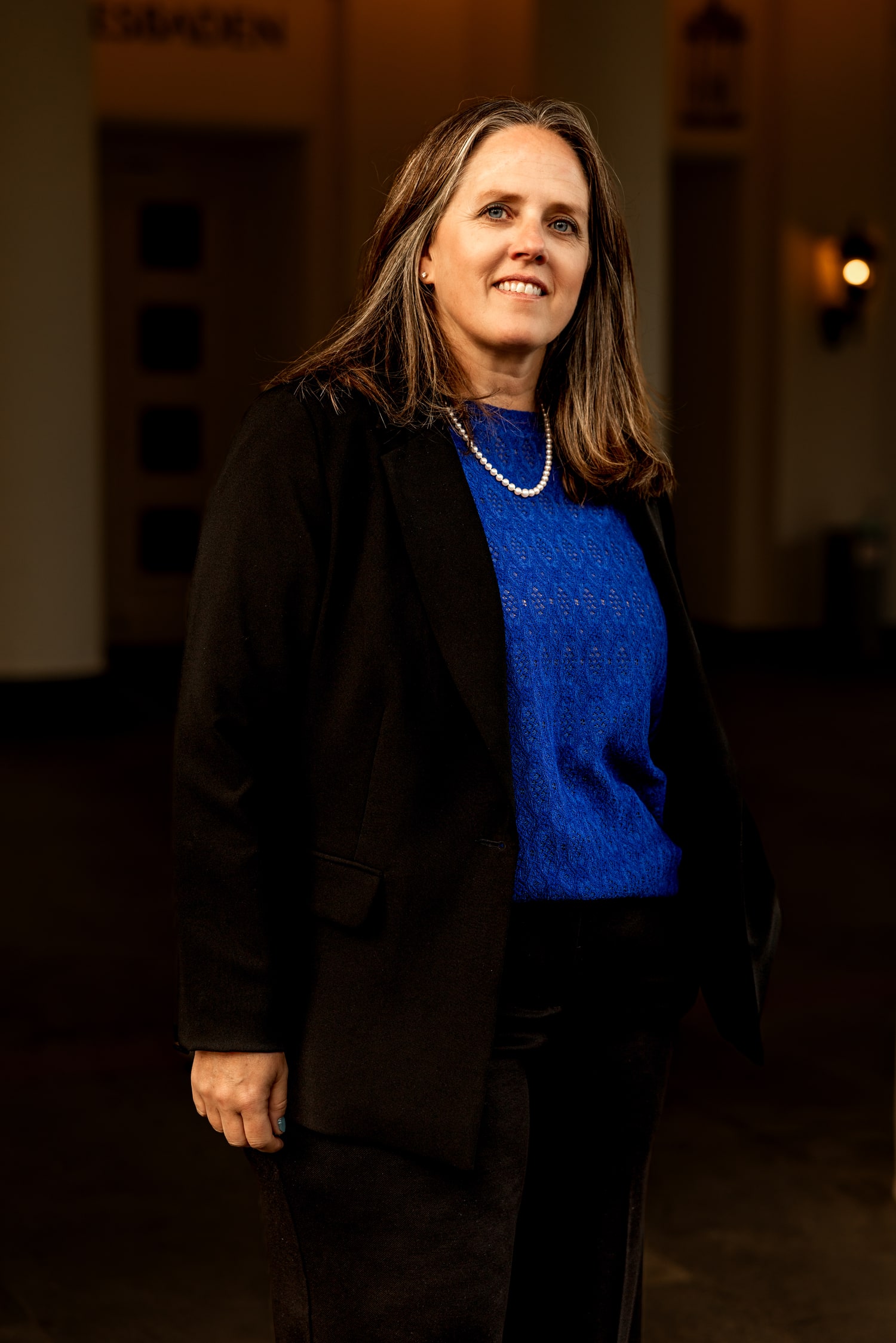 Kyle Meisner dressed in a dark blazer and necklace, standing in a softly lit hallway
