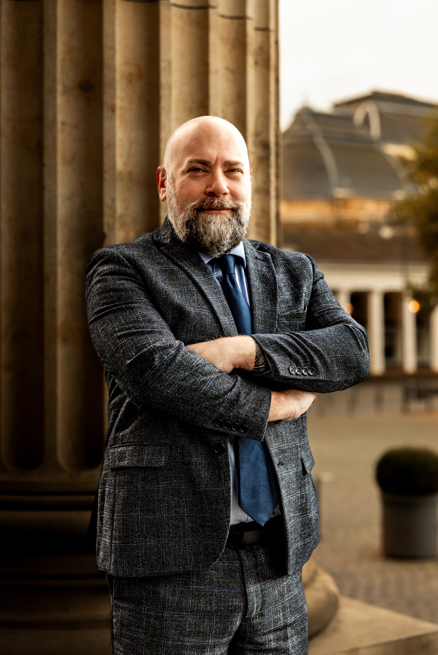 Matthew Krause in a checkered suit and blue tie, standing with arms crossed by stone columns outdoors