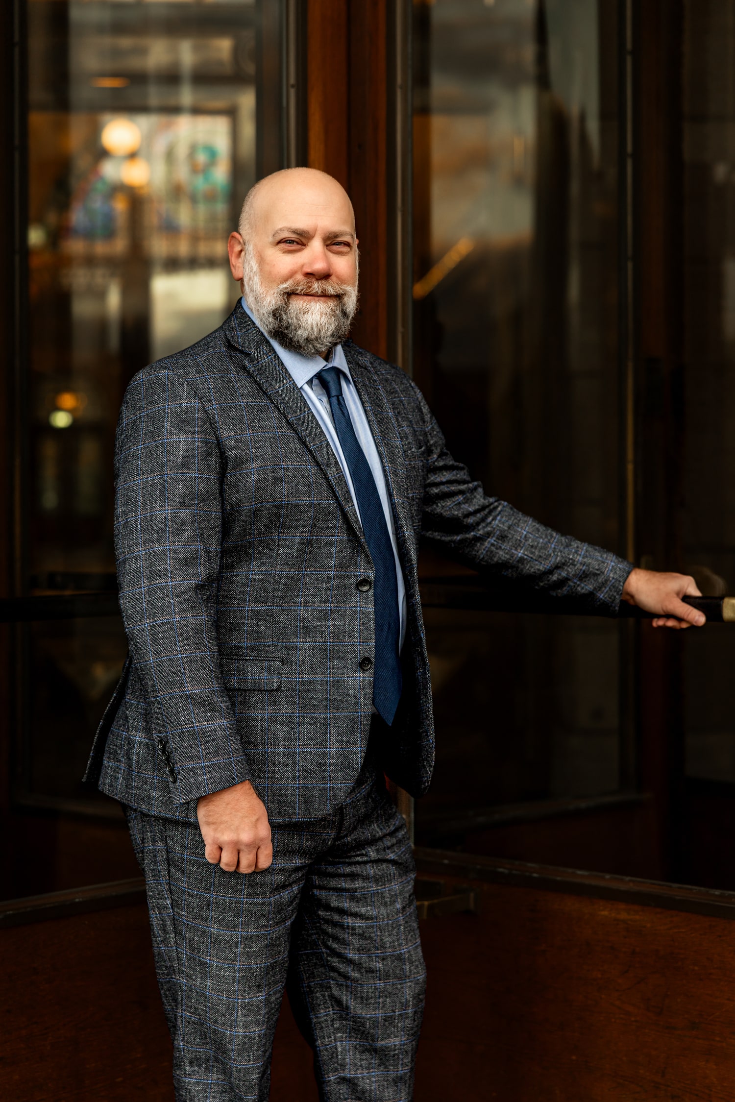 Matthew Krause dressed in a checkered suit and blue tie, holding open a wooden door