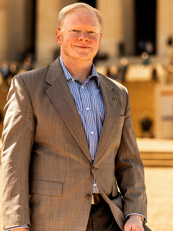 Military attorney Cadman Kiker standing outdoors, wearing striped shirt and tan suit jacket with building in background.