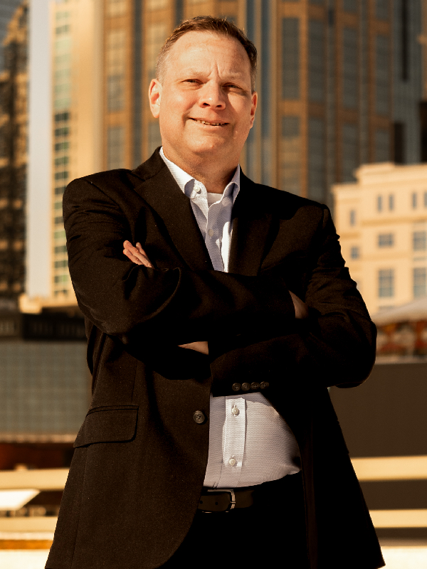 Military attorney EJ Gladding standing outdoors with arms crossed, wearing black suit and light blue shirt, city buildings in background