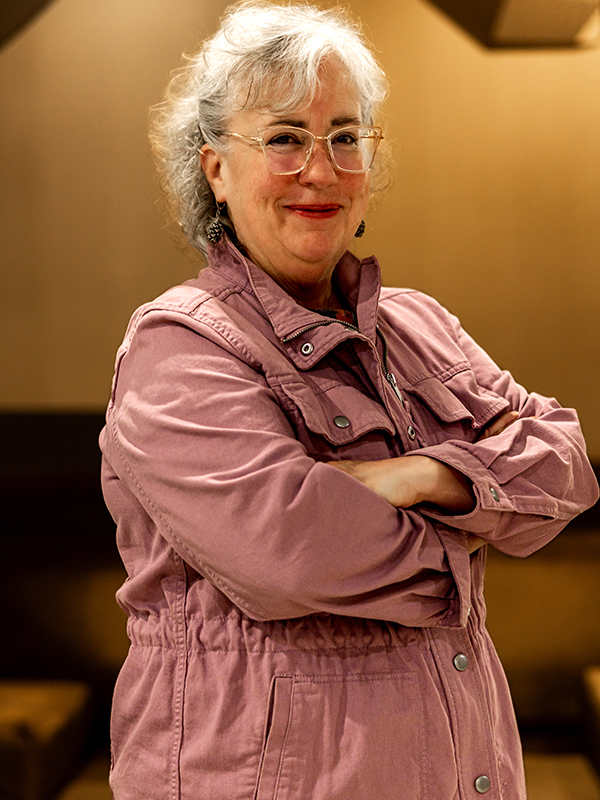 Military attorney Genie Hughes standing with arms crossed, wearing pink jacket indoors