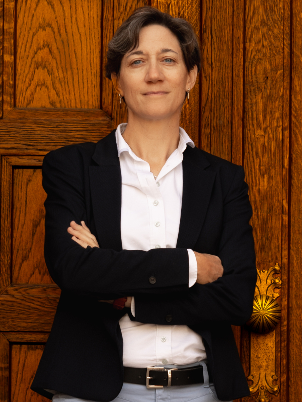 Military attorney Mary Ritzmann standing with arms crossed, wearing white shirt and black blazer in front of wooden door