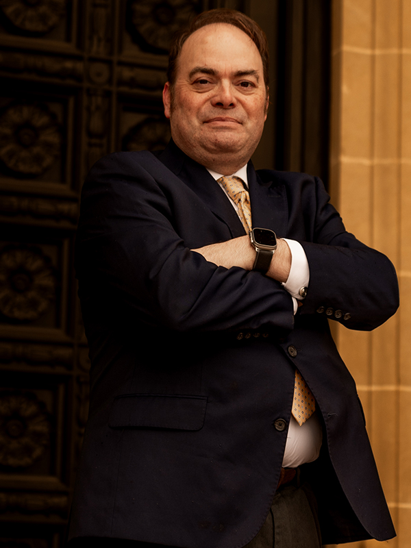 Military attorney Robert Newark standing outdoors, arms crossed, wearing navy suit and yellow patterned tie