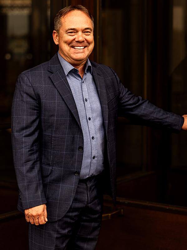 Military attorney Scott Brokaw standing indoors, wearing blue plaid suit and blue shirt, smiling