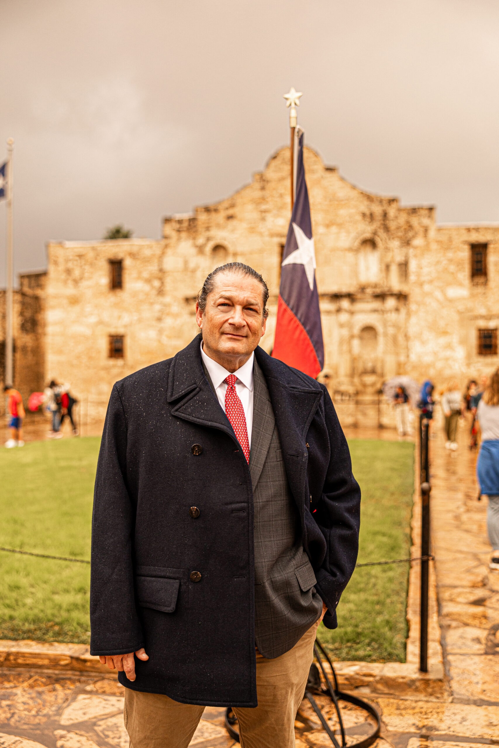 Will M. Helixon, Founder of the Law Office of Will M. Helixon, in a navy coat and red tie standing in front of the Alamo